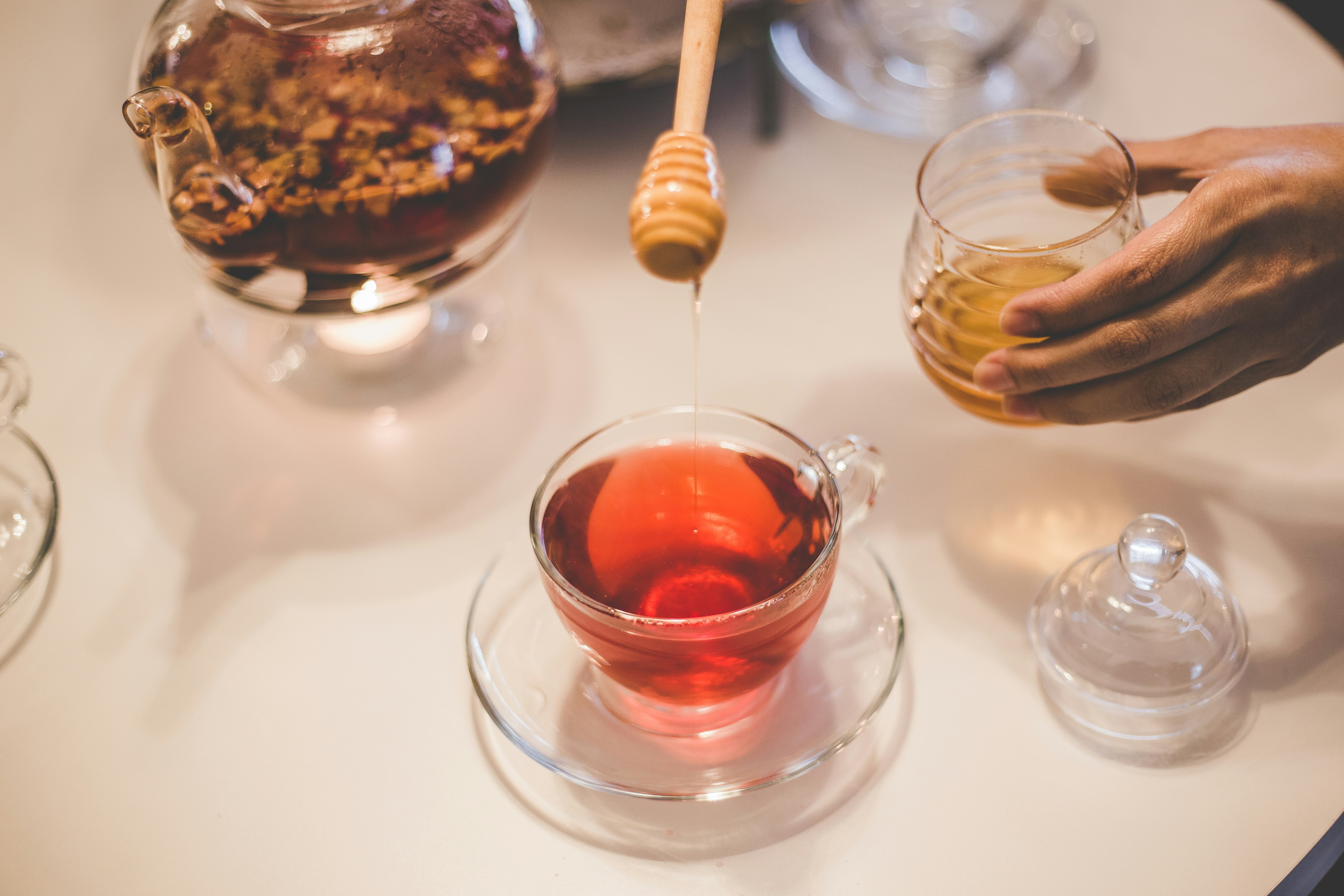 Person drizzling honey into tea cup with herbal tea, glass teapot on warmer in background